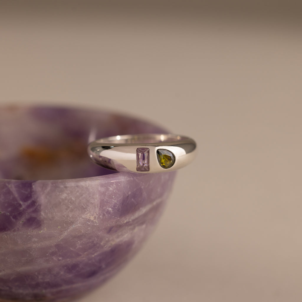 The Toi et Moi Dome Ring in Silver, featuring purple and yellow gemstones, rests elegantly on the edge of a purple marble bowl.