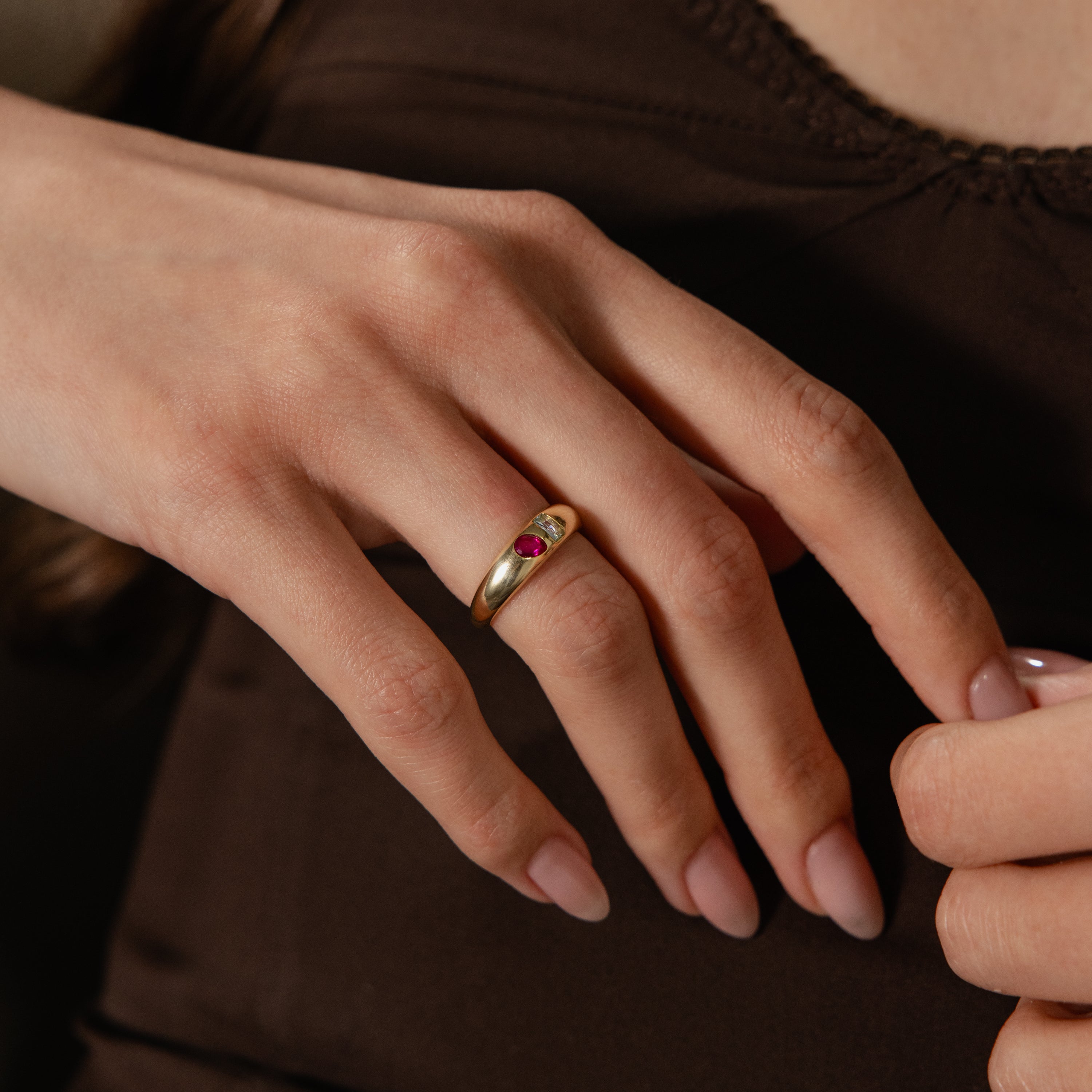 A hand with pale pink nails wears the Toi et Moi Dome Ring in 18K Gold, set with a small red gemstone and styled against a brown top.
