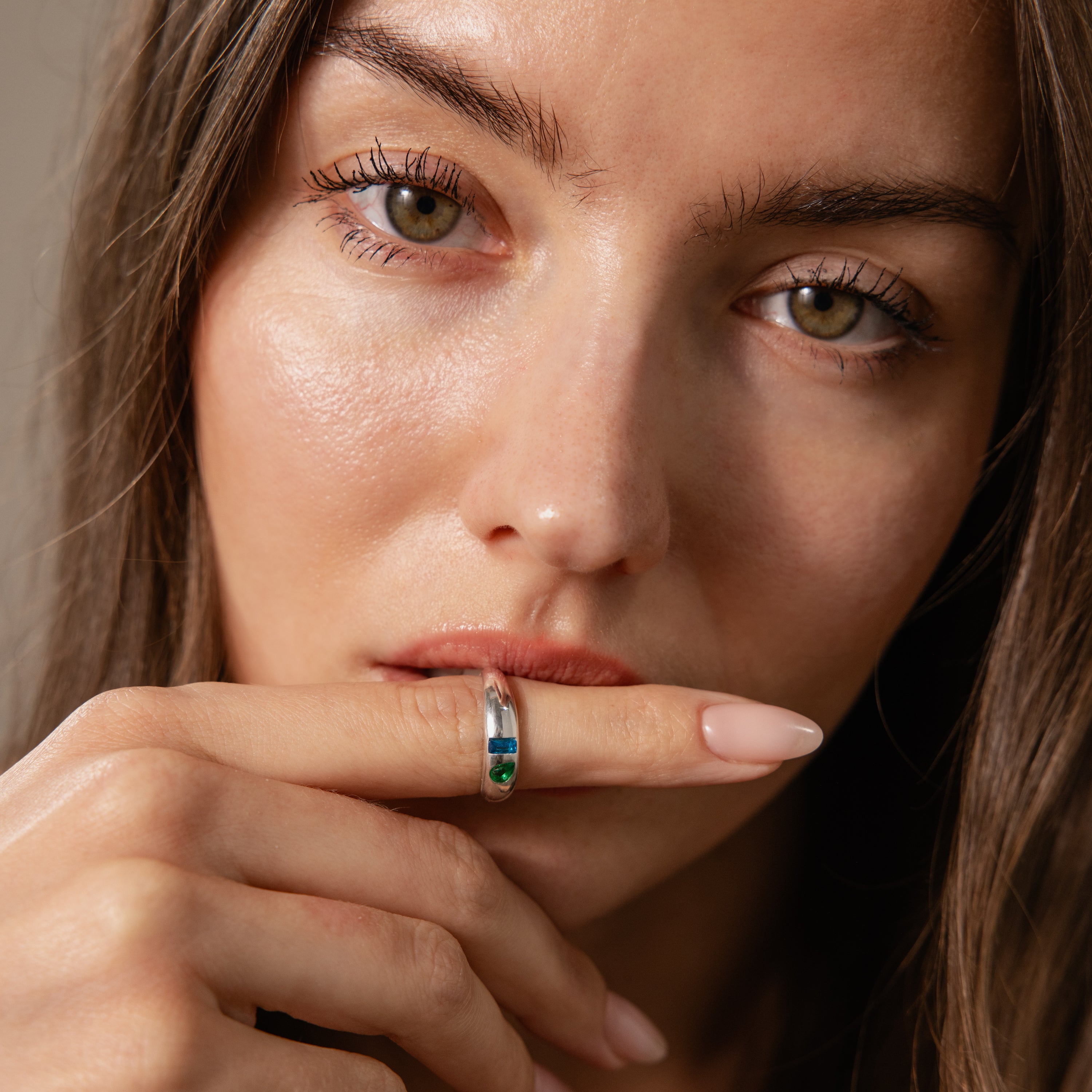 Woman with long brown hair touching her lips, wearing the Toi et Moi Dome Ring in Silver with green and blue stones on her finger.