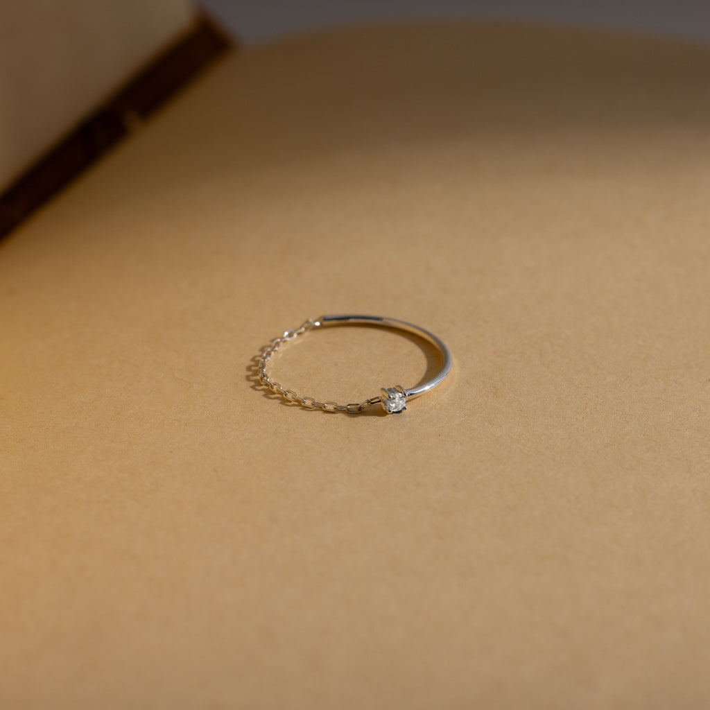 The Birthstone Half Chain Ring, a delicate silver piece with a small diamond, is beautifully displayed on a beige surface in soft lighting—perfect for lovers of dainty jewelry.