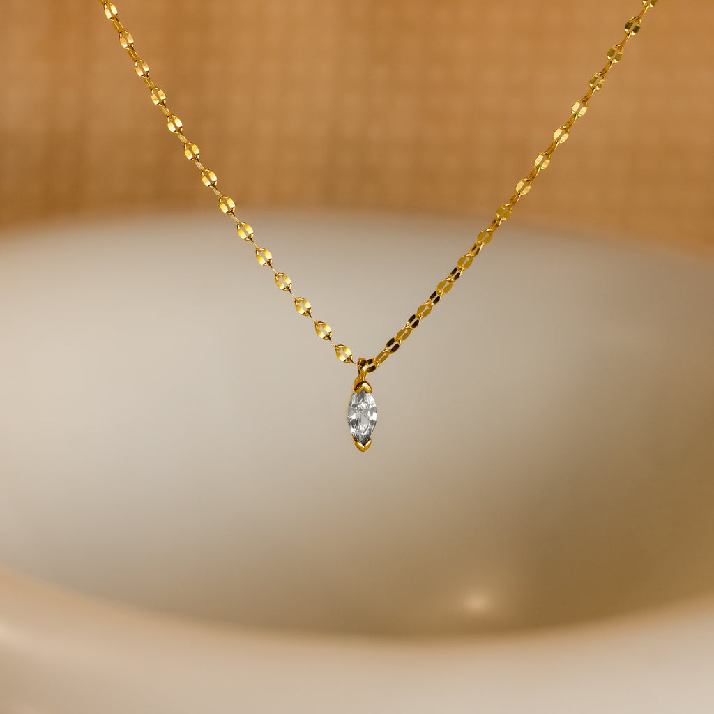 The Custom Marquise Birthstone Necklace is a gold chain with a marquise-cut diamond pendant, shown against a blurred beige background.