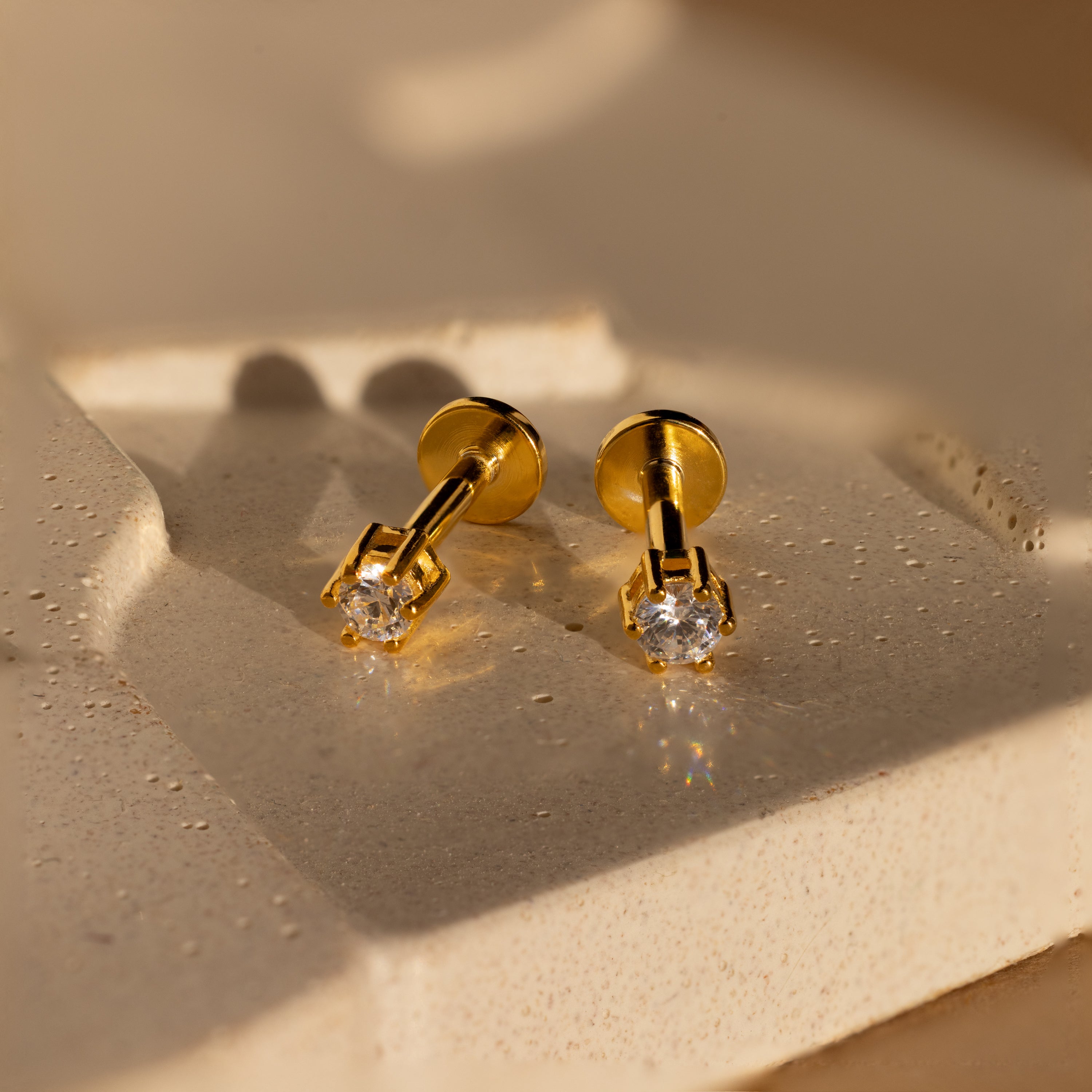 A pair of Diamond Flatback Studs with clear gemstones displayed on a textured beige surface.