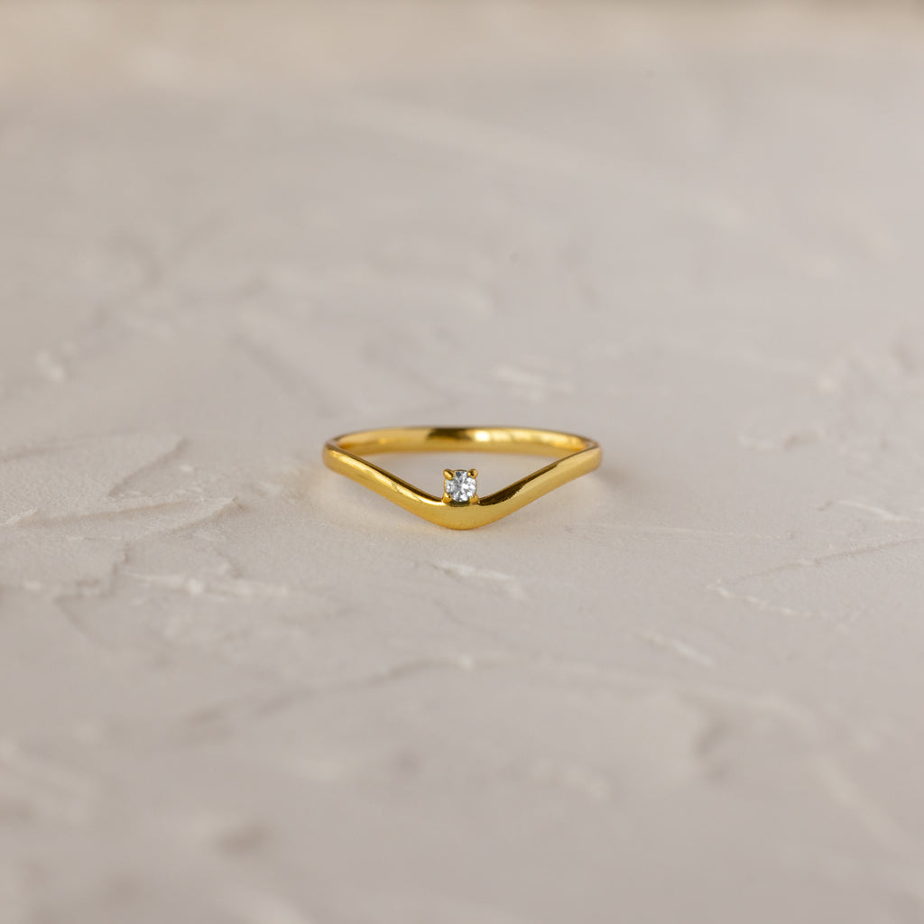 The Diamond Drop Curve Ring features a V-shaped gold band with a small central diamond, displayed on a textured light surface.
