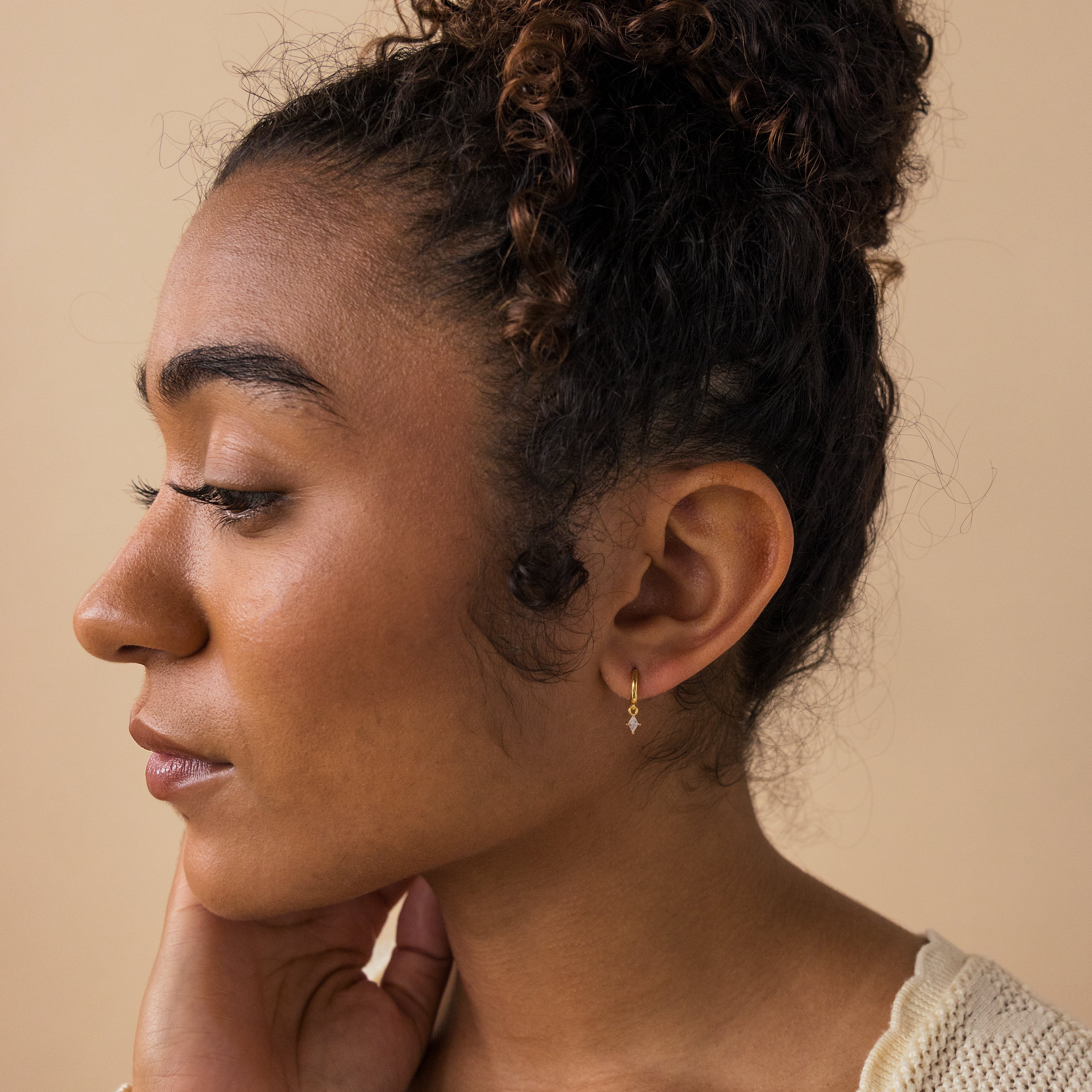 Woman with curly hair in a bun, wearing small gold Diamond Drop Huggies, looks to the left with a neutral expression.