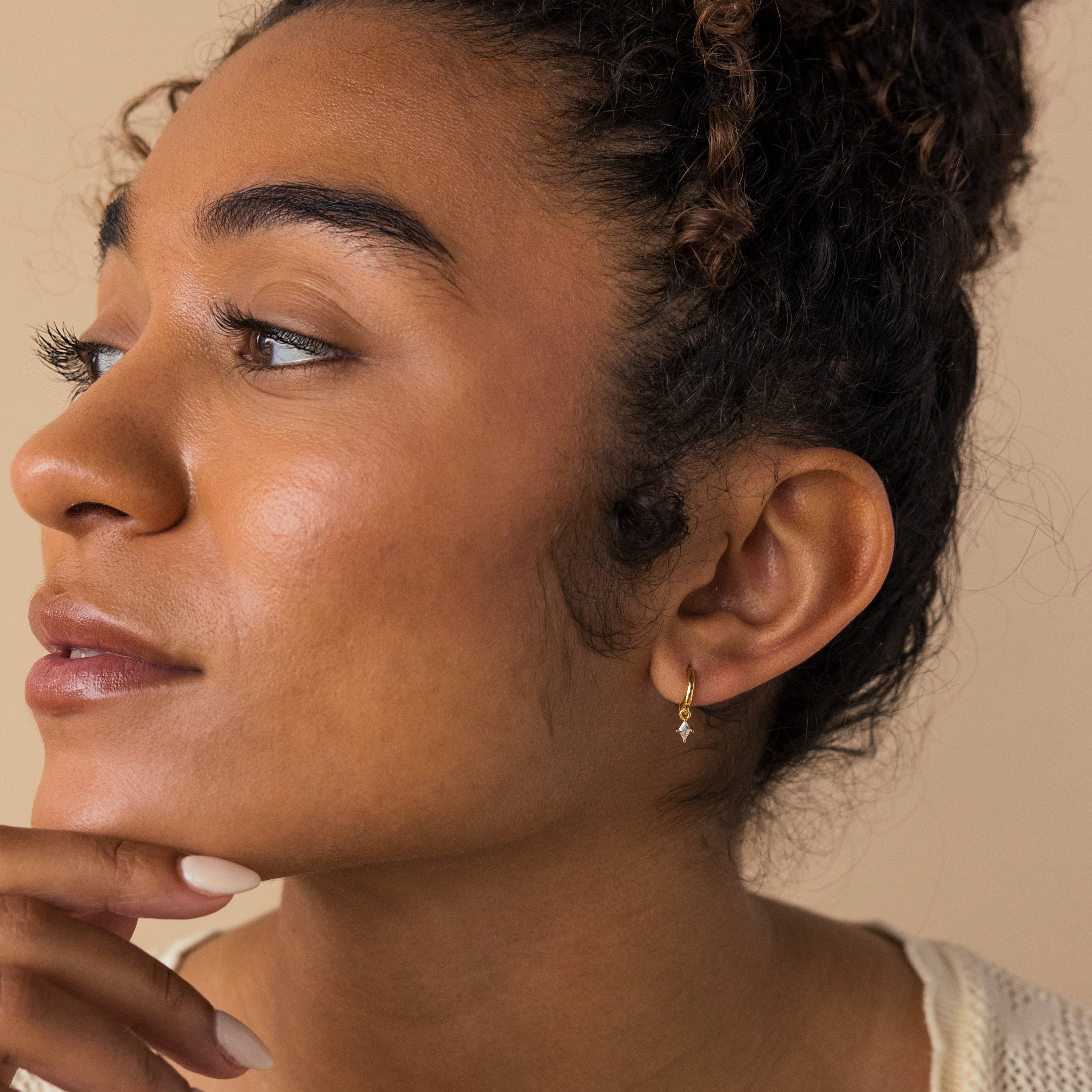 Woman with curly hair wears Birthstone Drop Huggies—a small gold earring—gazing left with her hand by her chin, showcasing the elegance of personalized jewelry.