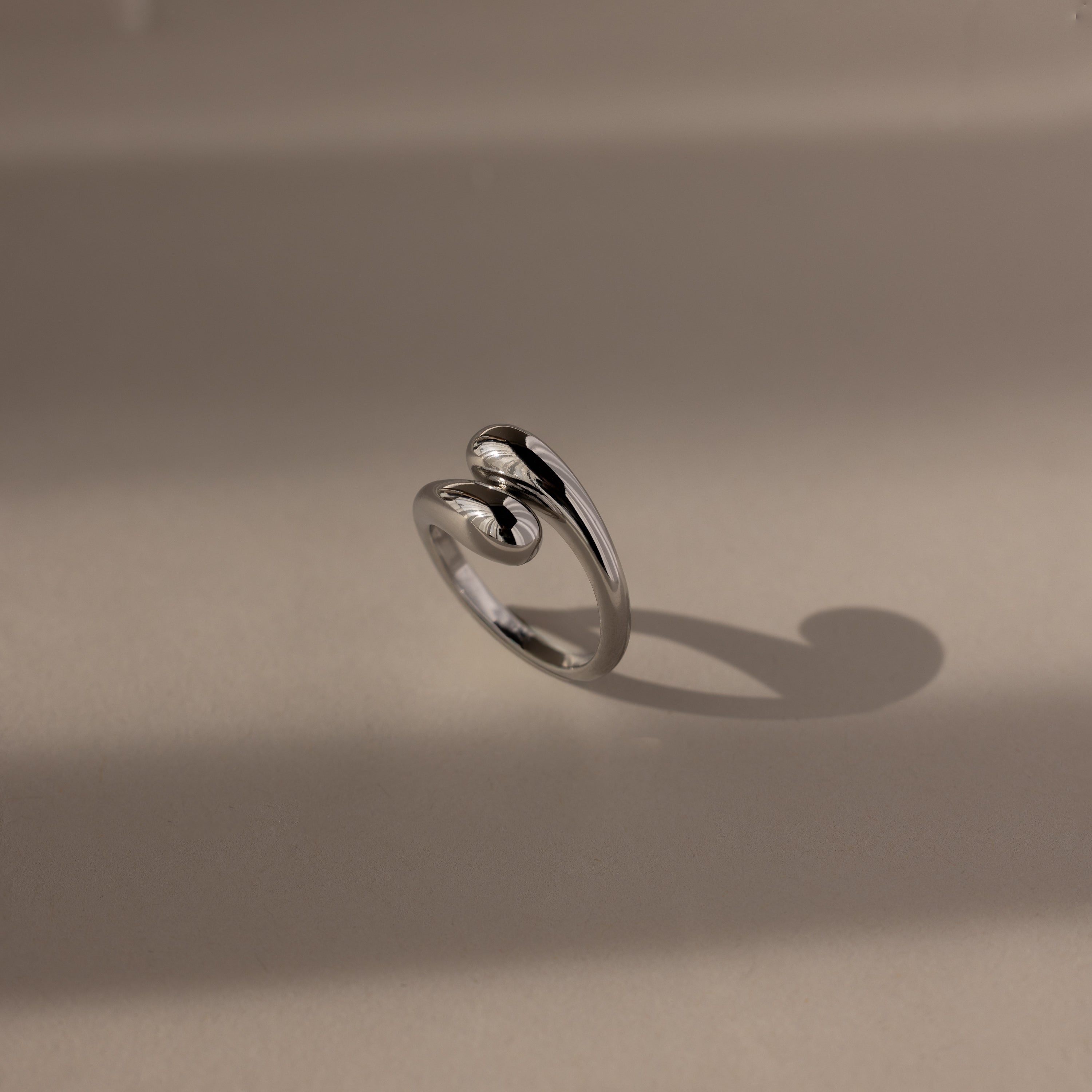 The Duo Dome Ring in White Gold features a modern, twisted design and casts a shadow on a beige surface—a sculptural jewelry piece with an adjustable band.