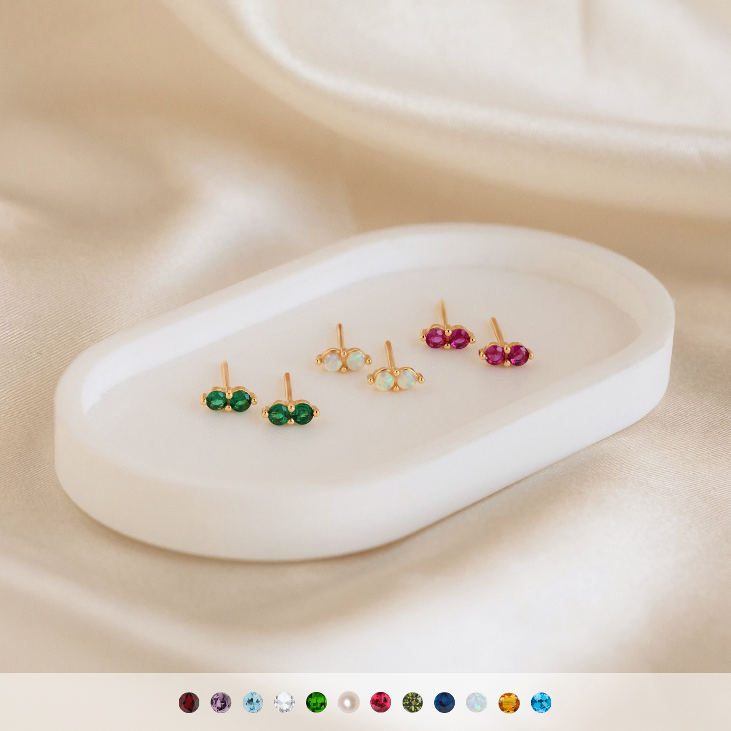 Three pairs of Duo Birthstone Studs in gold, featuring green, white, and pink stones, are displayed on a white tray over satin—an elegant and thoughtful bridesmaids gift.