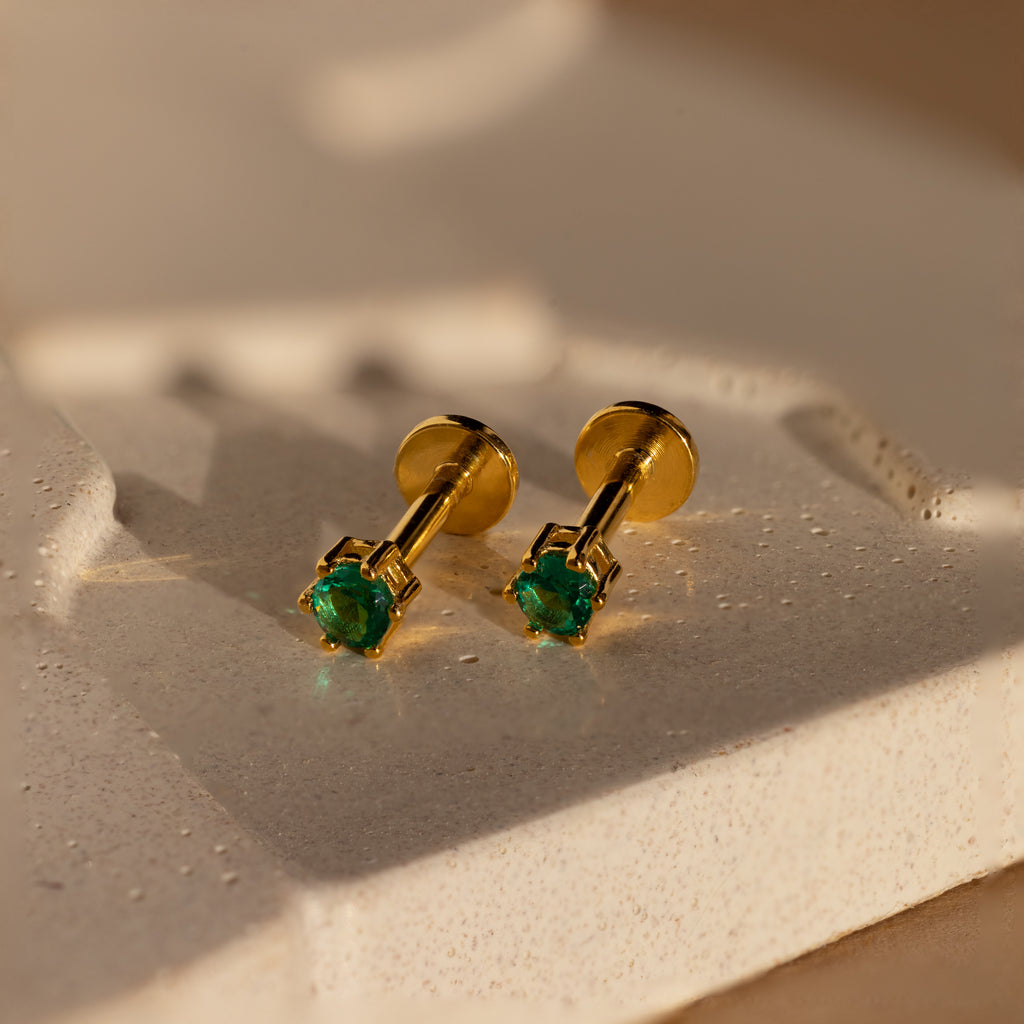 Emerald Flatback Studs: Elegant gold birthstone earrings with green gemstones, shown on a textured cream-colored surface.