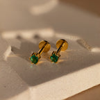 Emerald Flatback Studs: Elegant gold birthstone earrings with green gemstones, shown on a textured cream-colored surface.