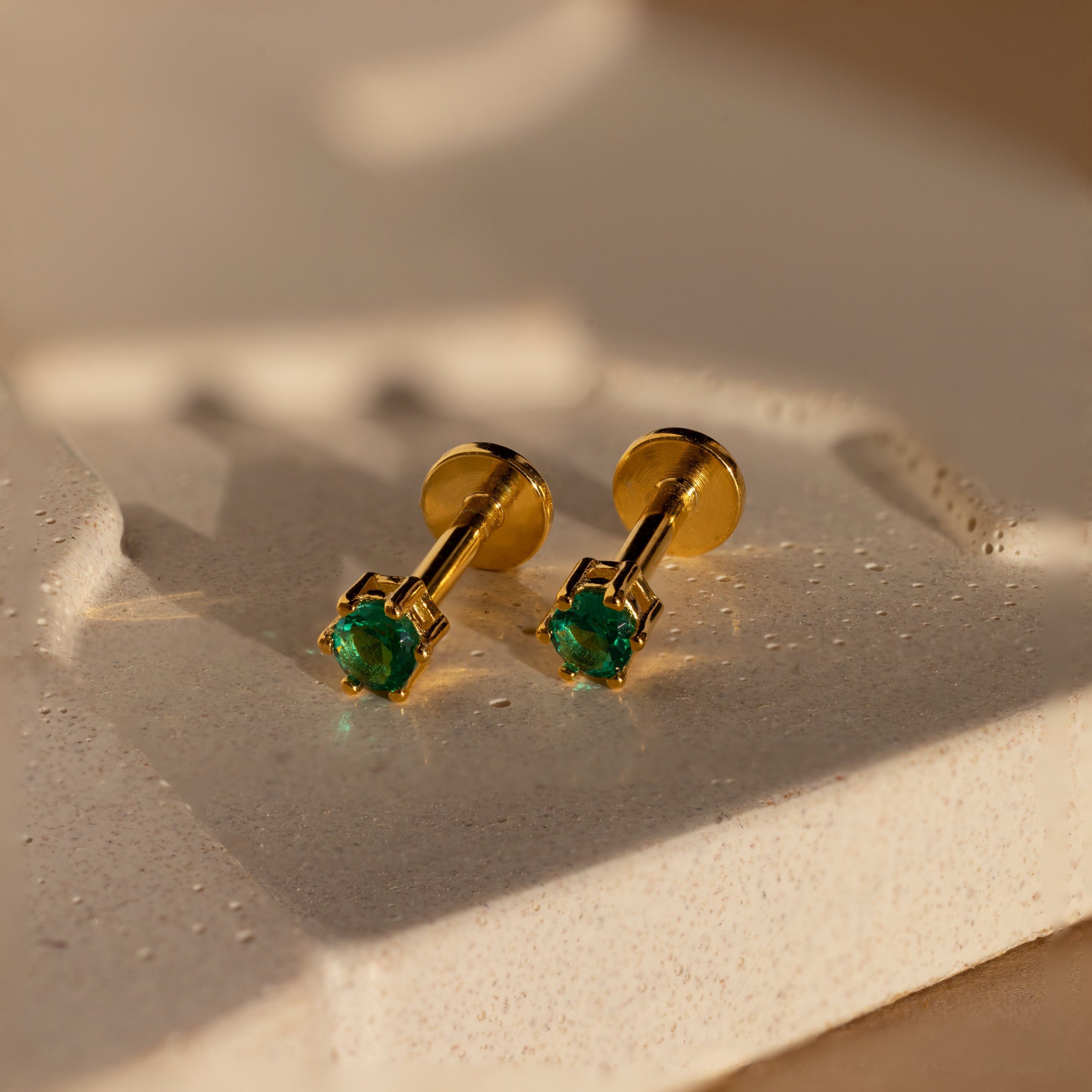 Emerald Flatback Studs: Elegant gold birthstone earrings with green gemstones, shown on a textured cream-colored surface.