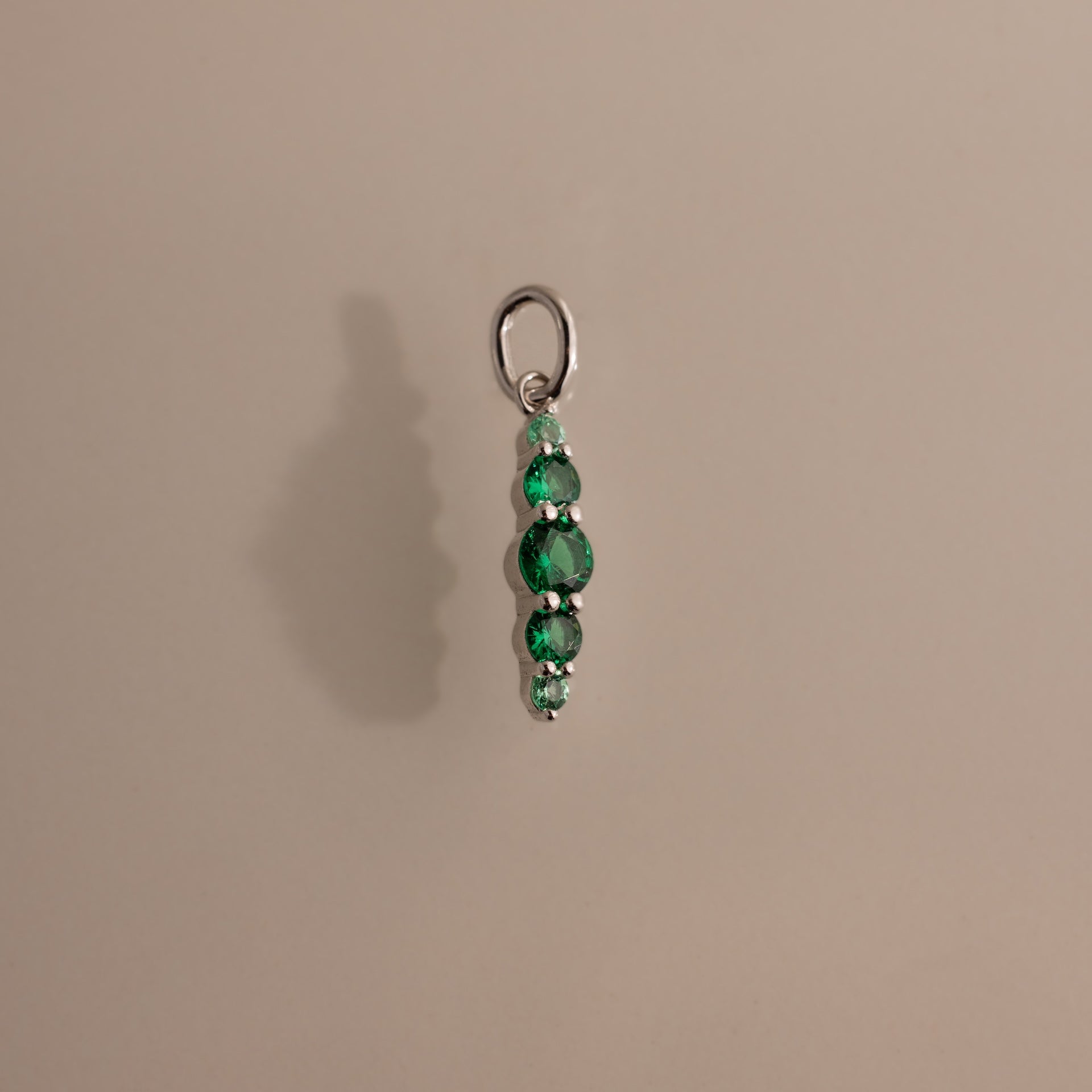 The Ombre Lace Emerald Charm features a silver pendant with four vertically stacked green beads, casting a faint shadow on beige—an elegant birthstone addition to any collection.