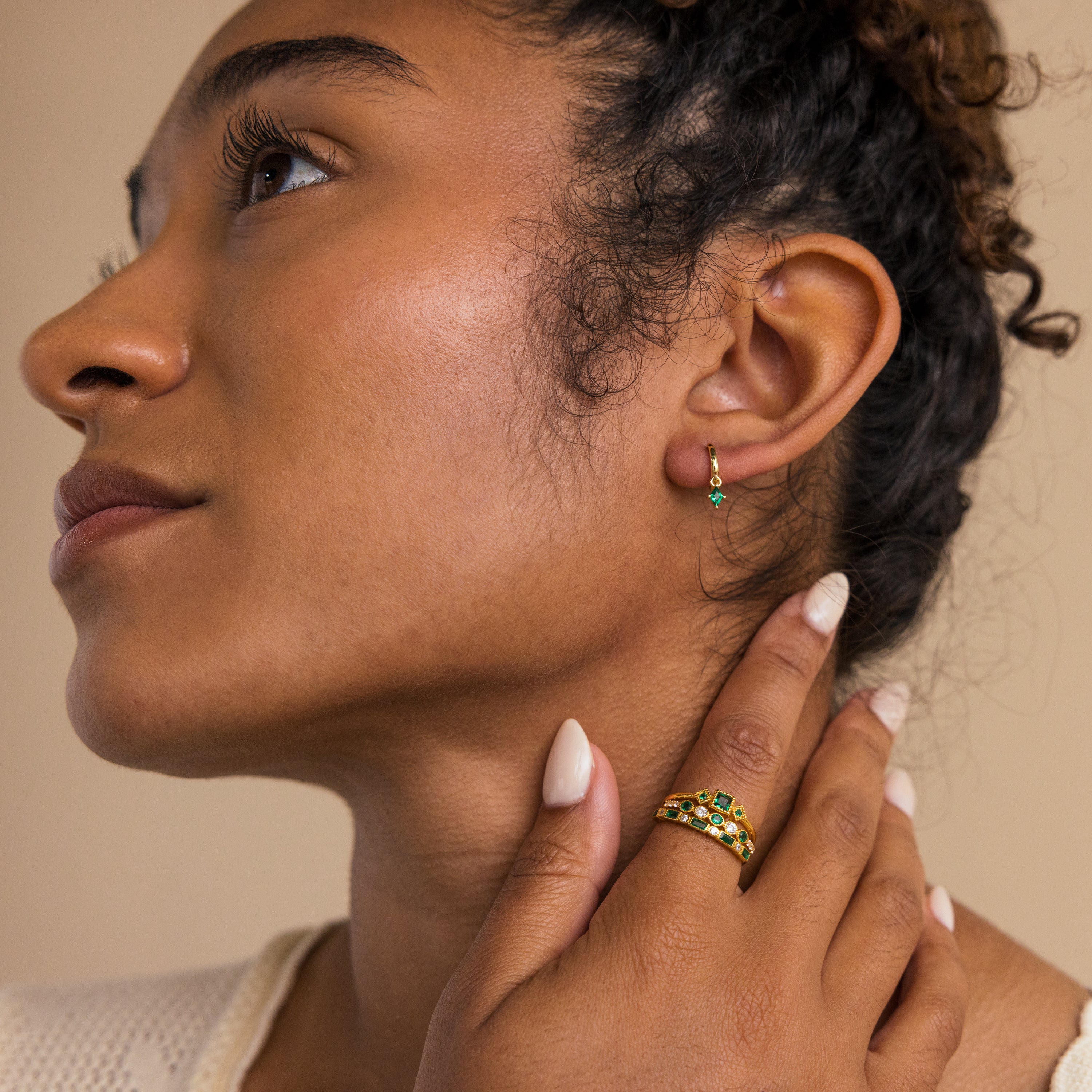 A woman with curly hair wears Emerald Drop Huggies and gold rings with green stones, touching her face gently to showcase personalized jewelry.