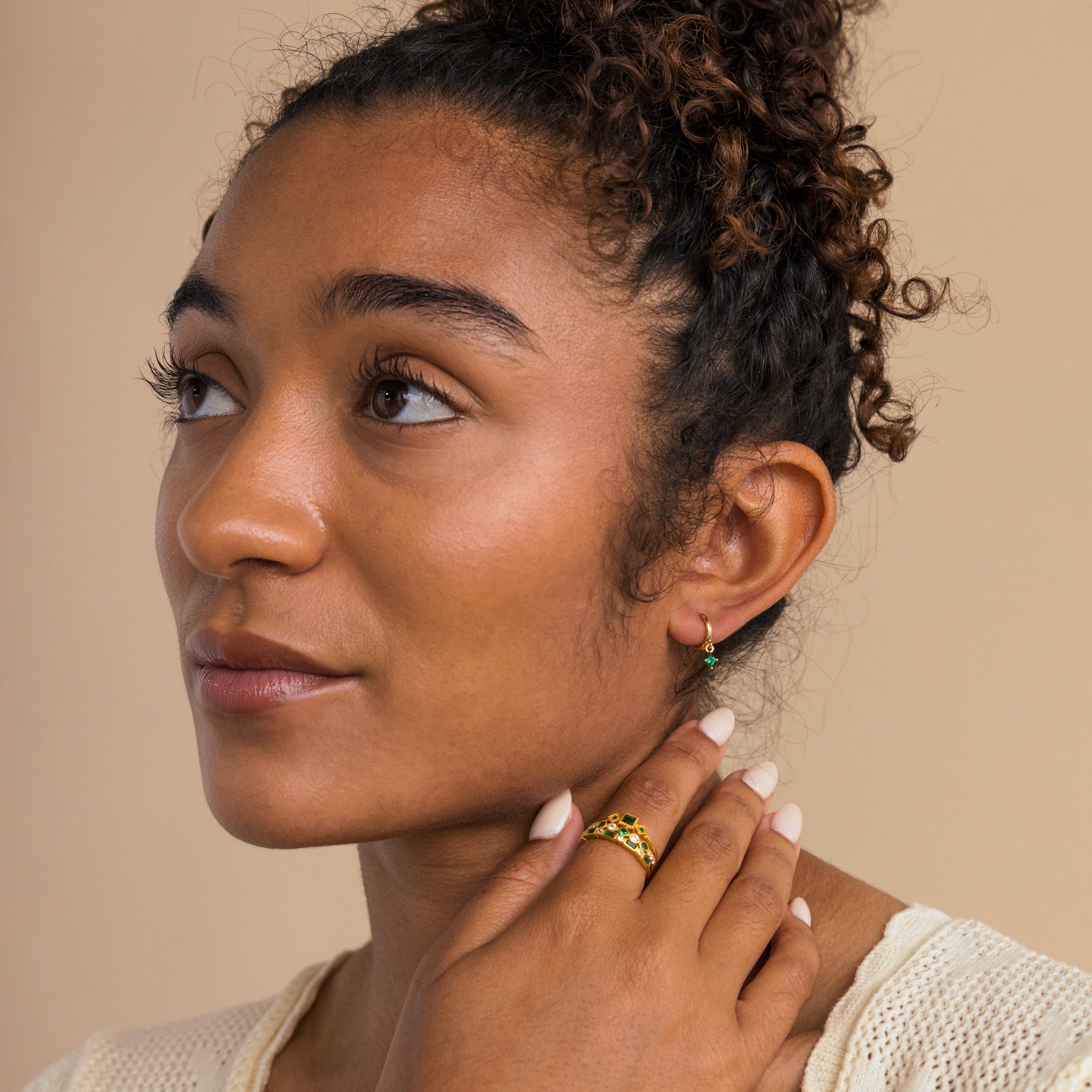A woman with curly hair wears Birthstone Drop Huggies and a gold ring, gazing to the left against a neutral background.