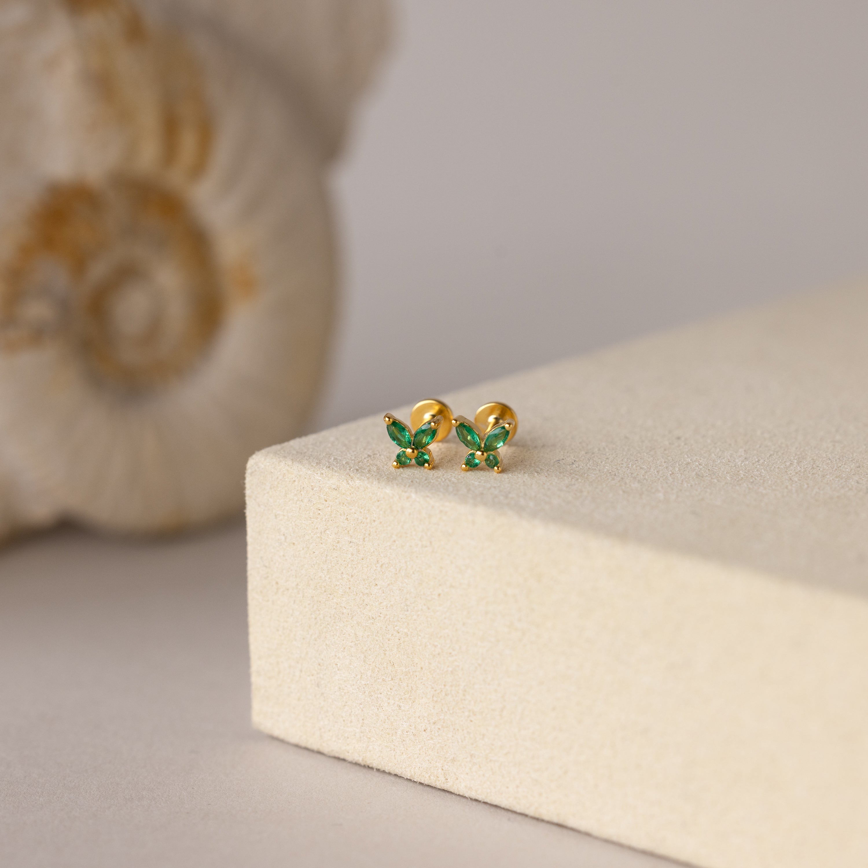 The Butterfly Emerald Flatback Studs—gold earrings with small green gemstones—add a personalized jewelry touch, shown on a beige platform beside a shell.