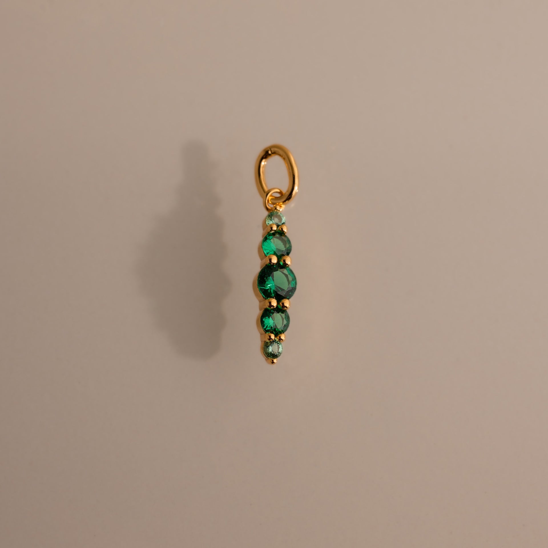 A gold Ombre Lace Emerald Charm with four stacked green beads on a plain beige background.