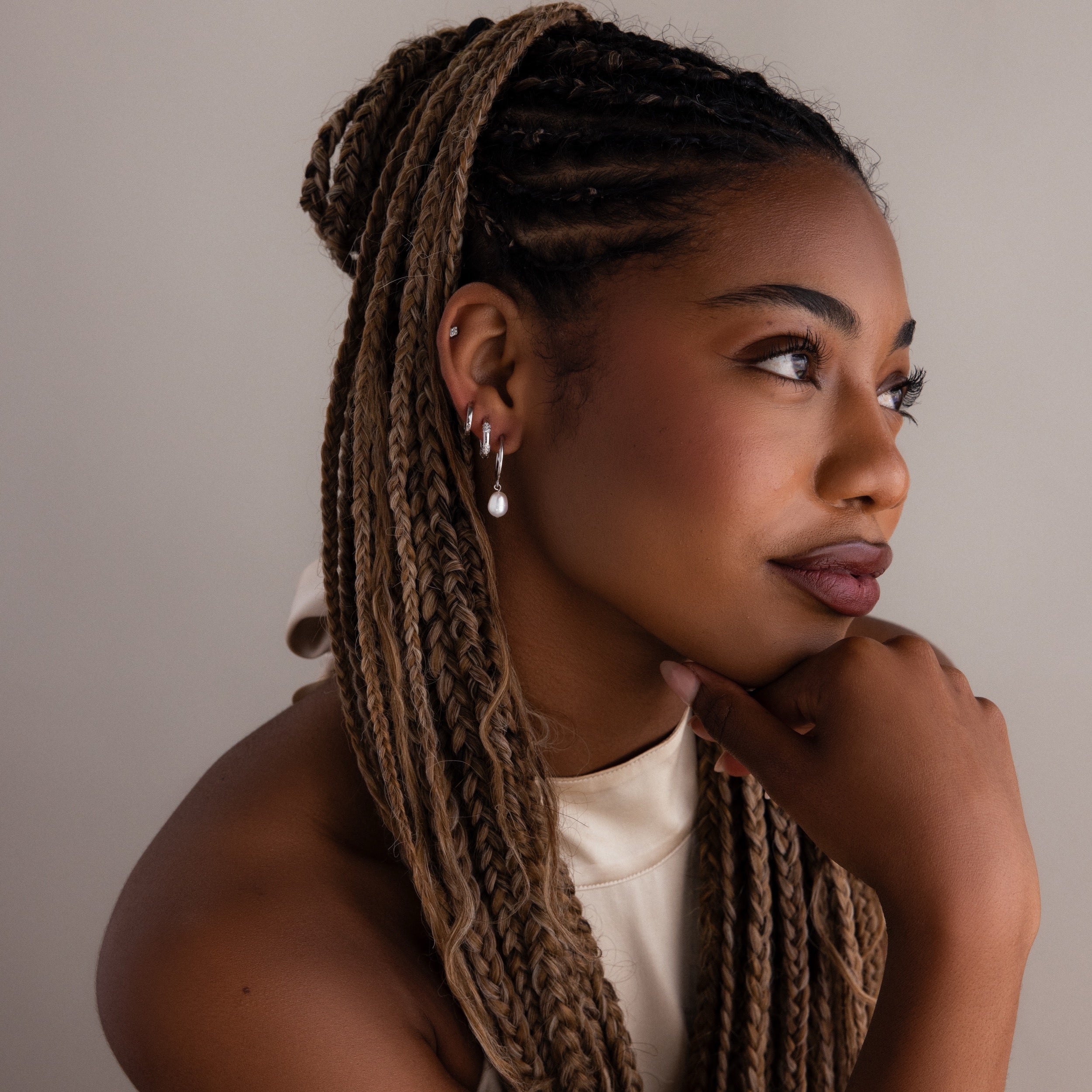 Woman with braided hair wearing various silver earrings and a white top against a neutral background