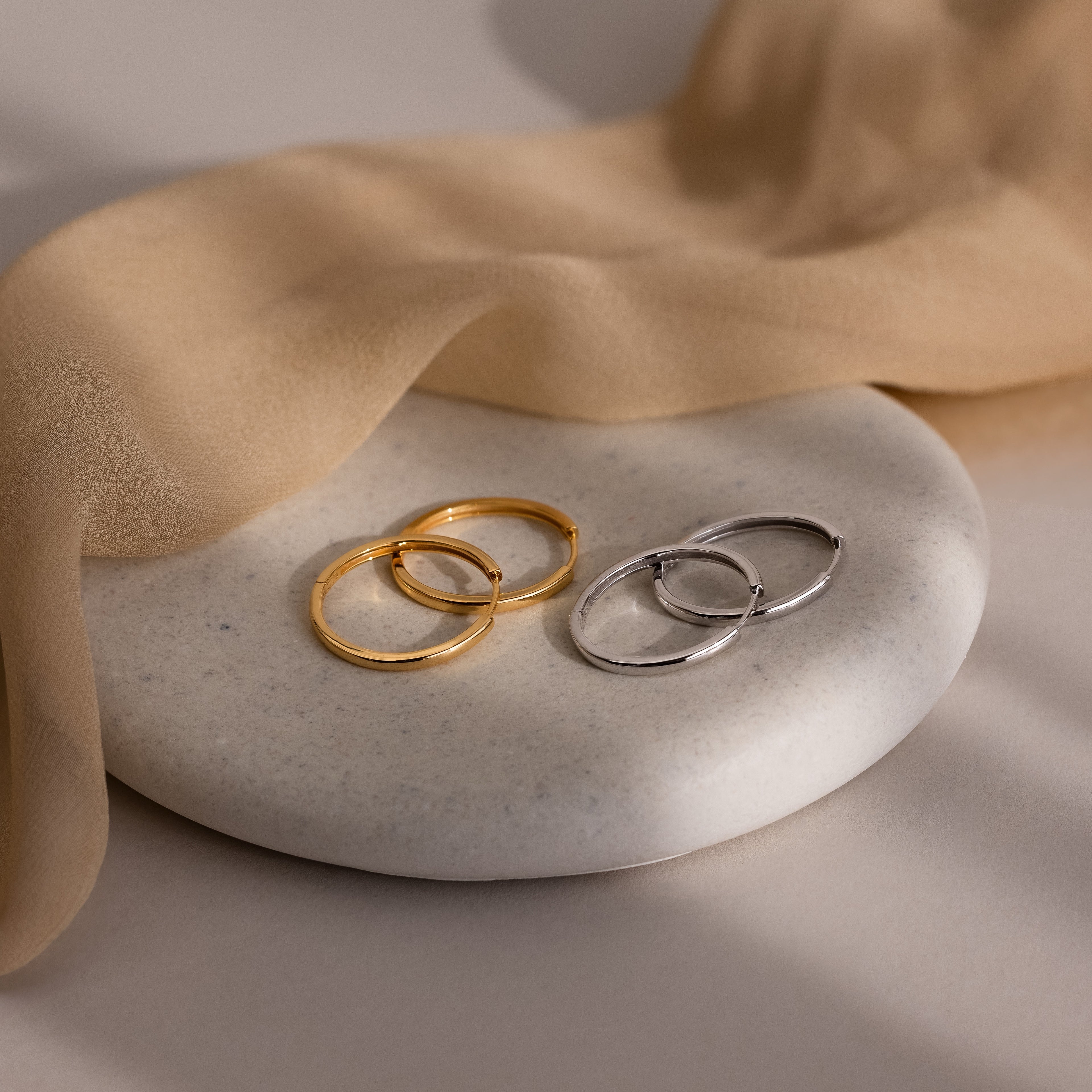 Two pairs of gold and silver hoop earrings on a stone surface with beige fabric.