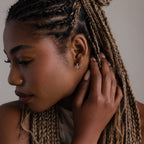 Woman with braided hair touching her ear, wearing a small diamond hoop earring.