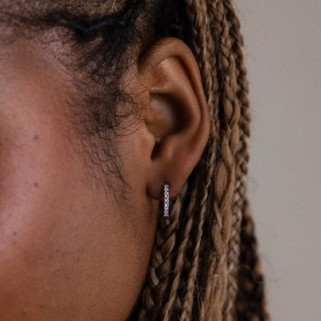 Close-up of an ear with braided hair wearing a small pave hoop earring against a neutral background