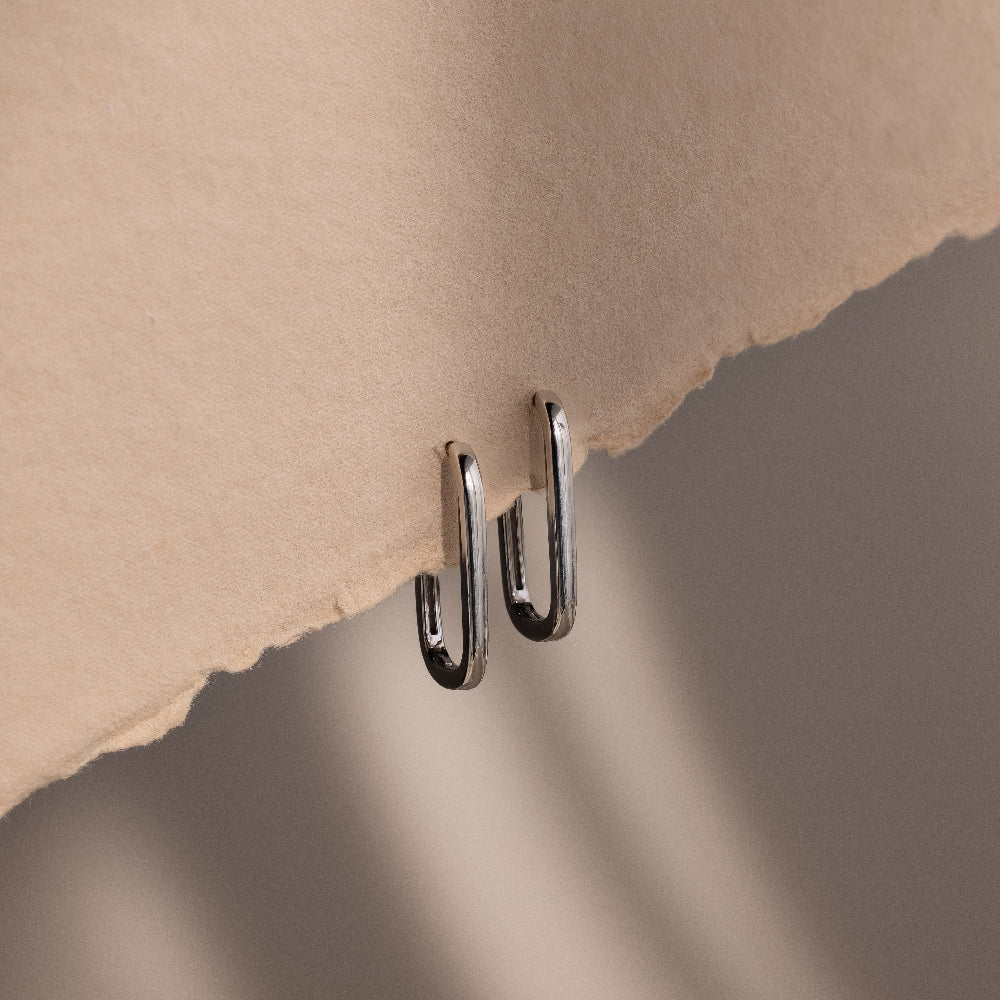 Metallic silver rectangular hoop earrings attached to a piece of beige paper against a gradient background