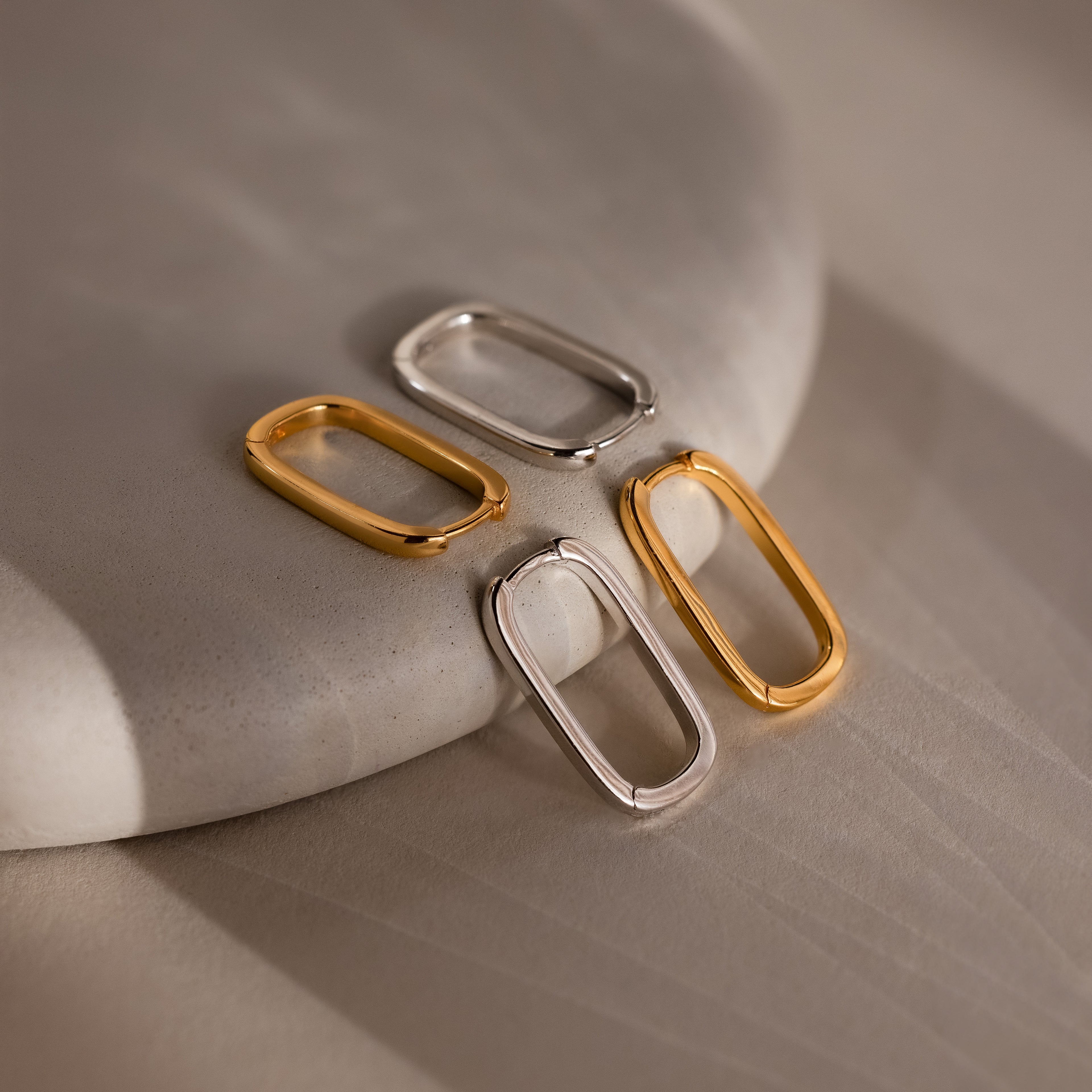 Four metallic silver and gold rectangular hoop earrings on a textured surface