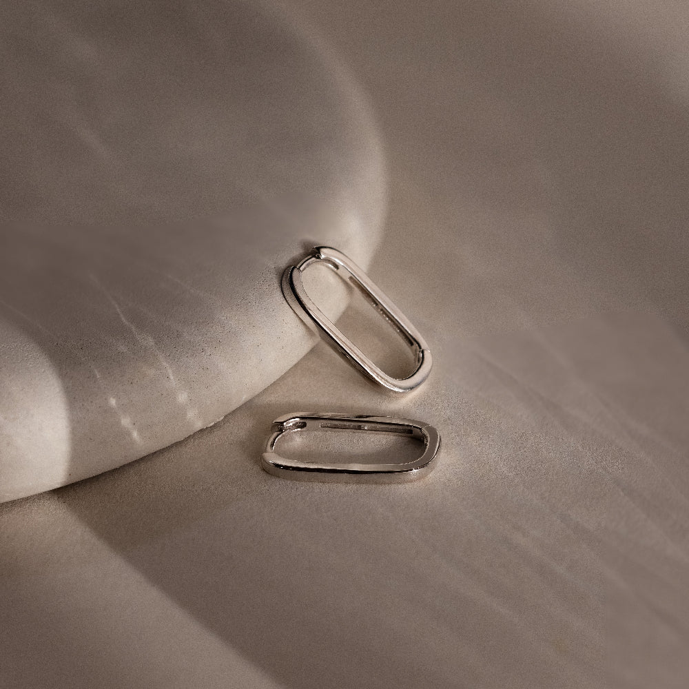 Two silver rectangular hoop earrings on a textured beige surface