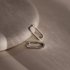 Two silver rectangular hoop earrings on a textured beige surface