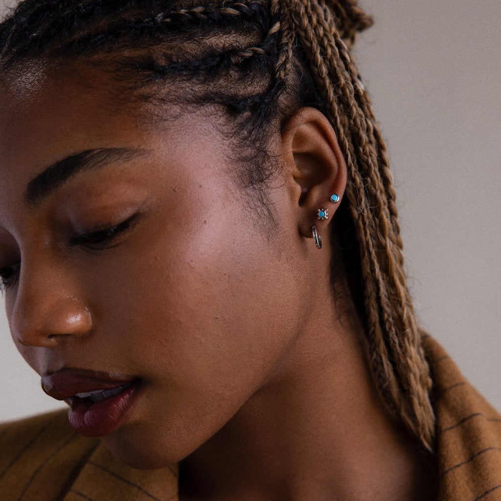 Close-up of a woman with braided hair wearing various silver turquoise earrings against a neutral background