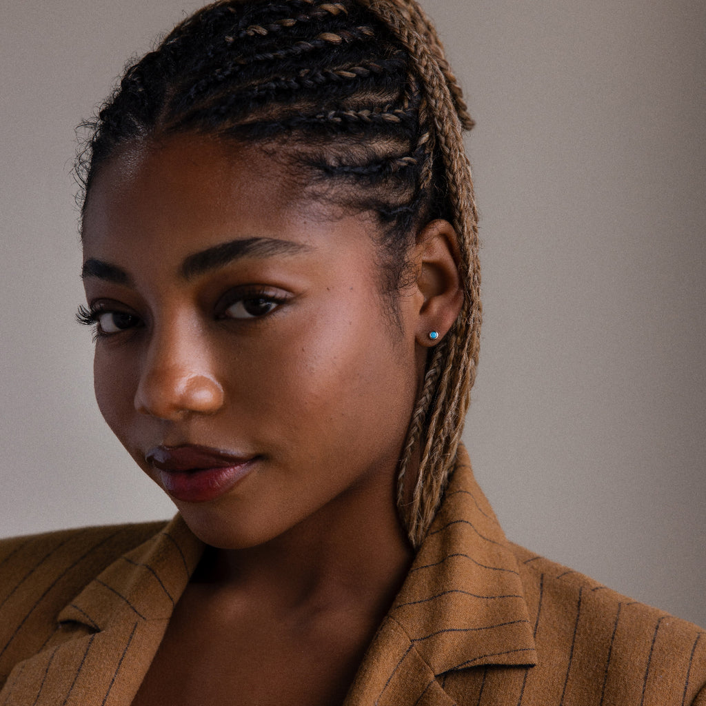 Woman with braided hair wearing a brown jacket and silver turquoise stud earrings against a neutral background