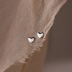 Heart-shaped silver stud earrings on a textured beige surface