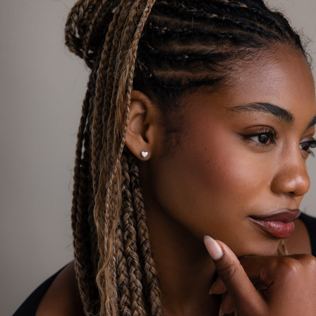 Woman with braided hair and a silver heart shaped stud earring touching her face against a neutral background