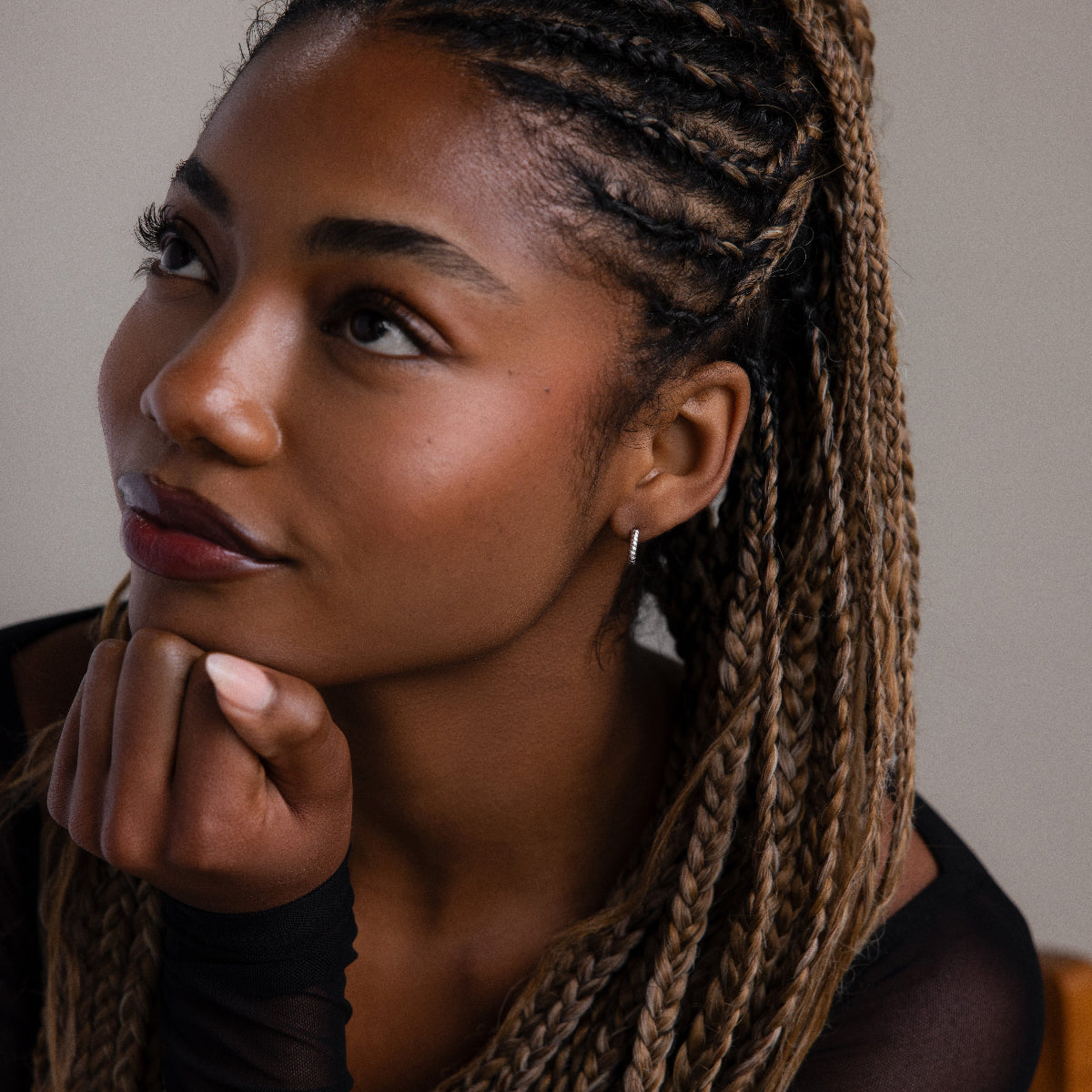 Woman with braided hair and a silver twisted hoop earring resting her chin on her hand against a neutral background