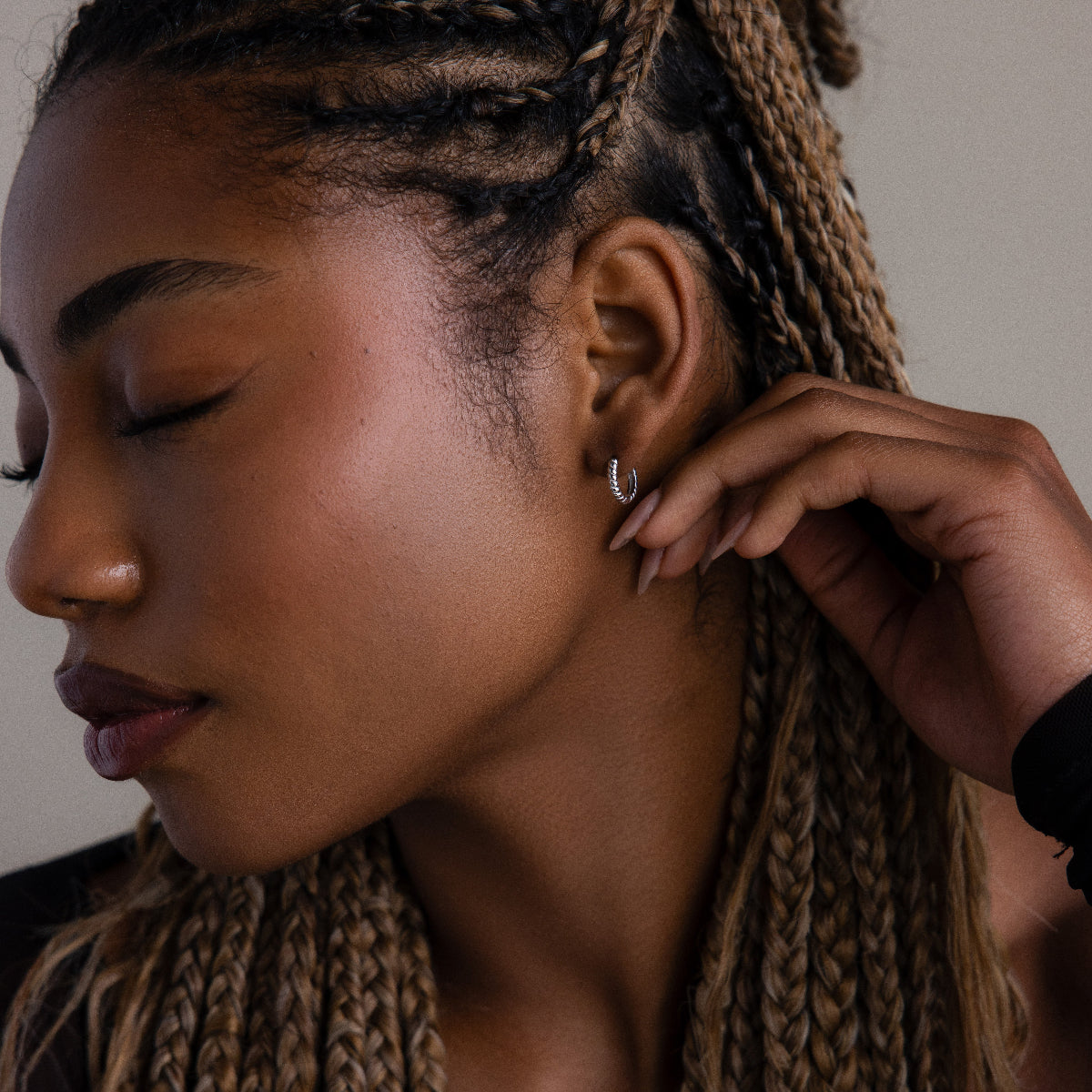 Woman with braided hair touching her ear, wearing a silver twisted hoop earring.
