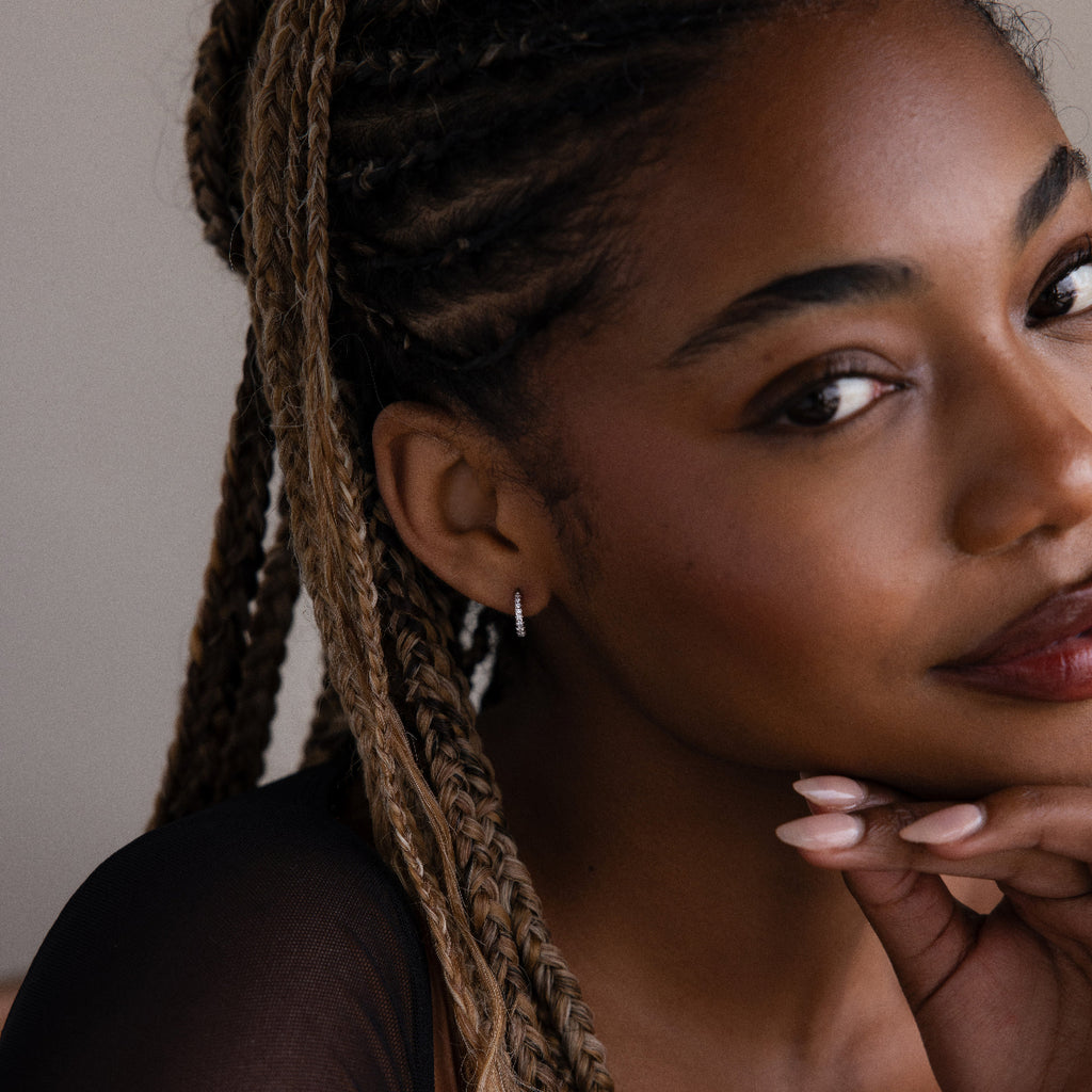 Woman with braided hair and a silver diamond hoop earring resting her head on her hand against a neutral background