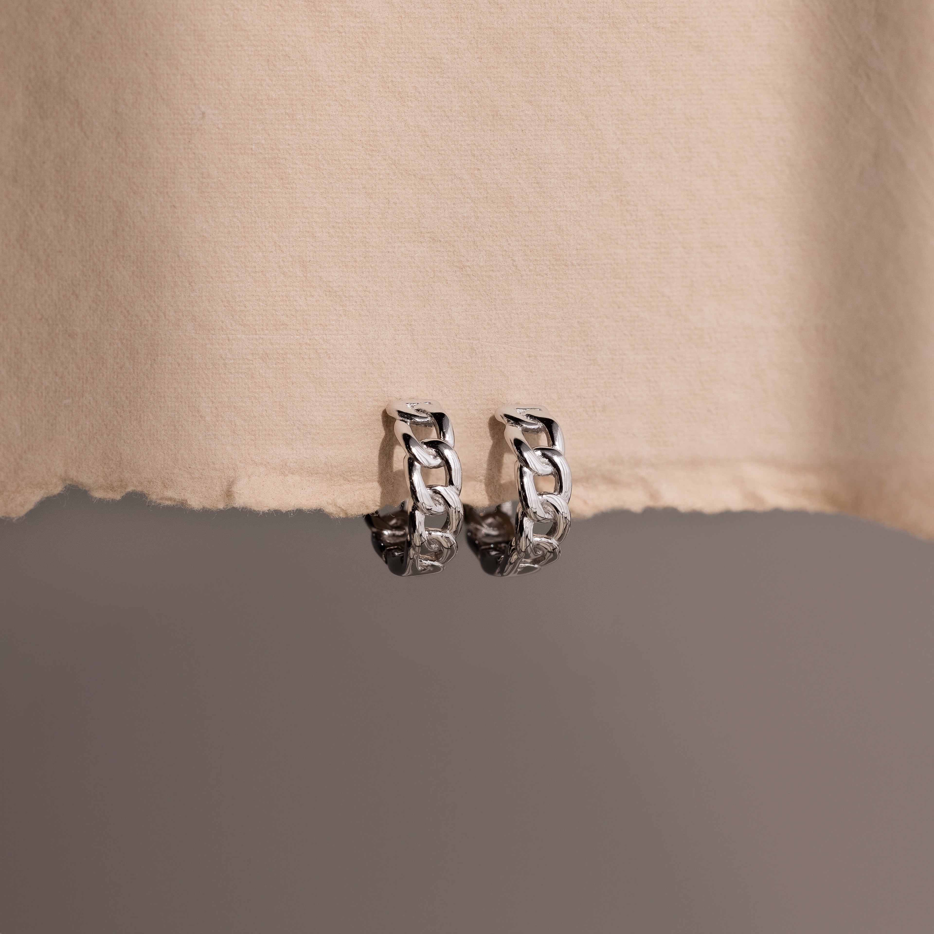 Pair of silver chain hoop earrings on a textured beige surface.