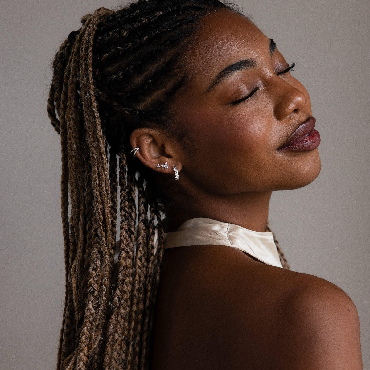 Woman with braided hair wearing multiple silver earrings leaning her head back against a neutral background