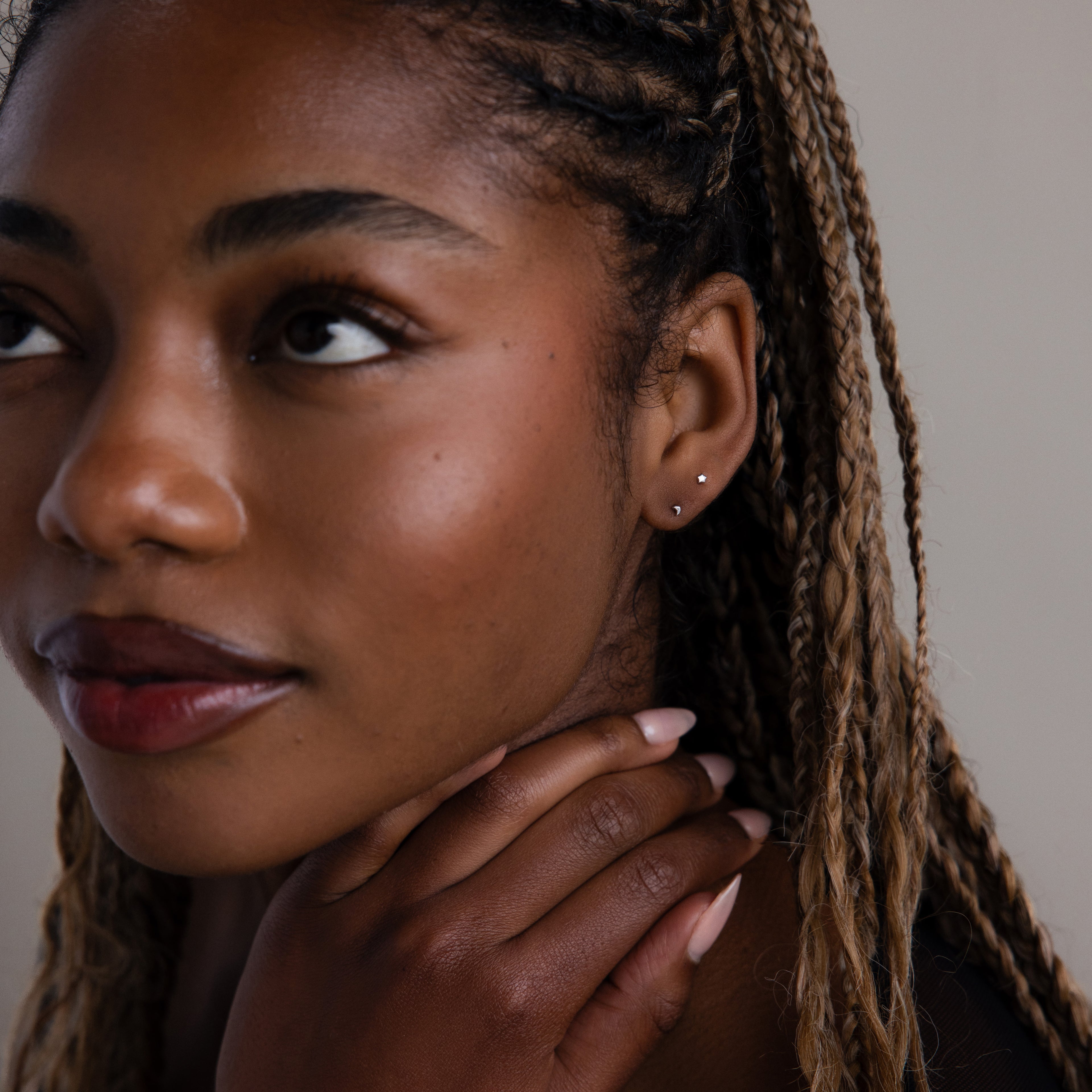 Woman with braided hair and tiny silver moon and star earrings touching her neck against a neutral background