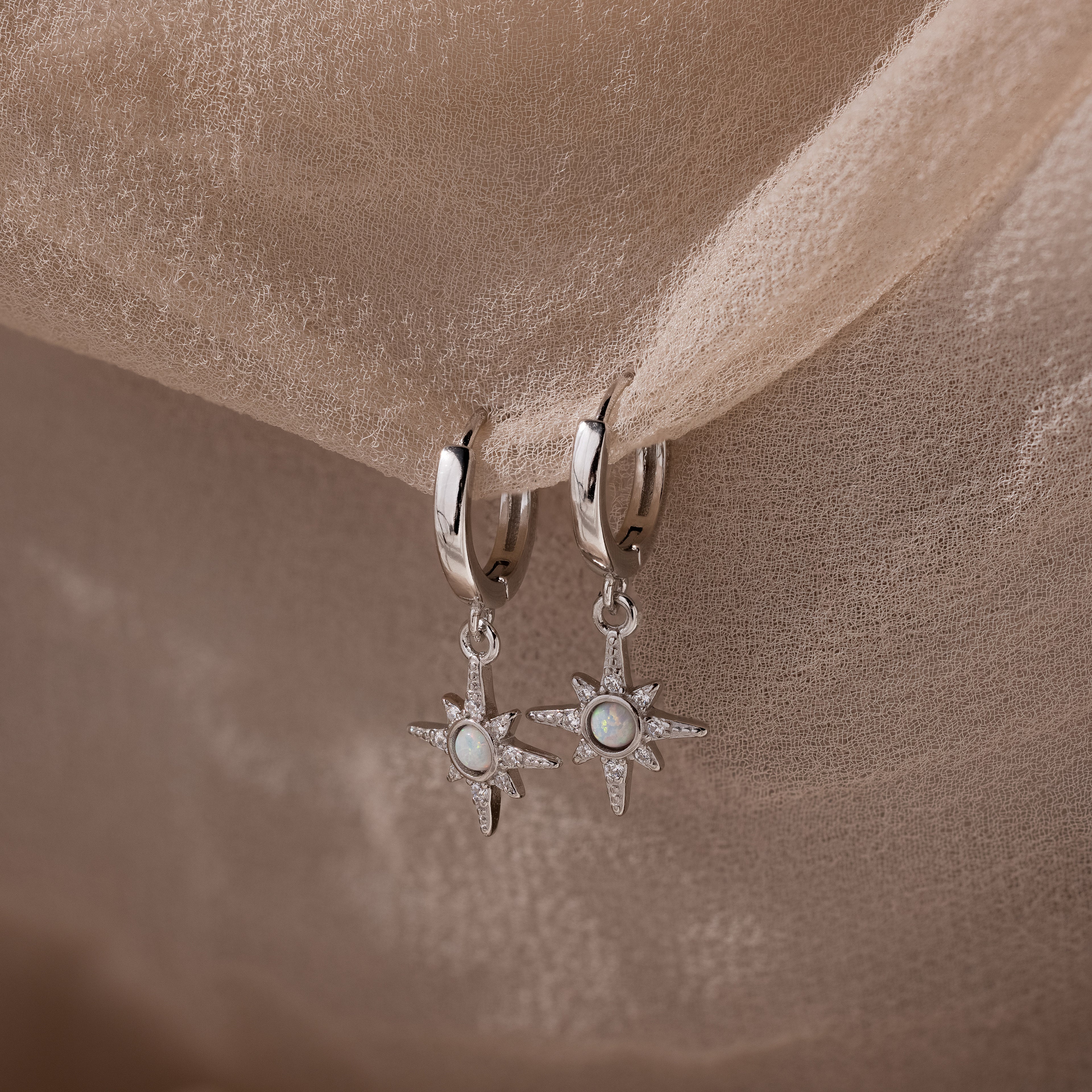 Pair of hanging silver opal hoop earrings with star designs on a beige fabric background