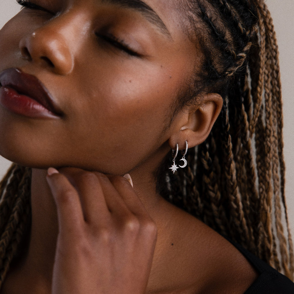 Close-up of a woman with braided hair wearing silver moon and star earrings, with a neutral background