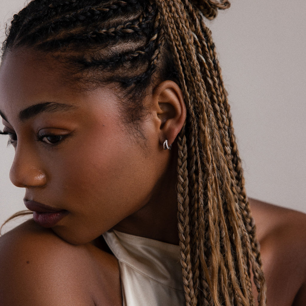 Woman with braided hair wearing silver hoop earrings and a white top against a neutral background