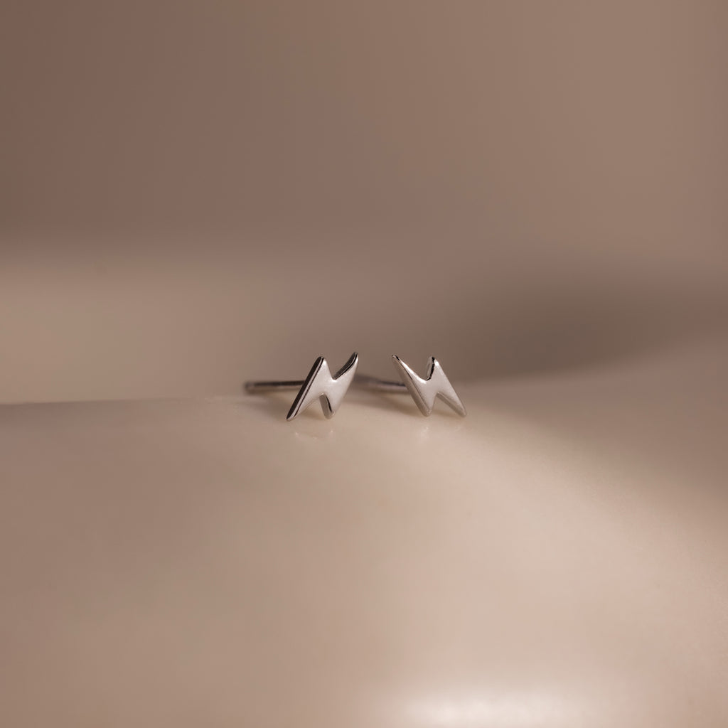 Close up of dainty silver lightning bolt stud earrings displayed on a soft neutral surface with minimal aesthetic styling.