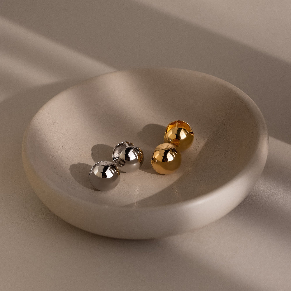 Four metallic sphere earrings in gold and silver on a beige ceramic dish with a neutral background