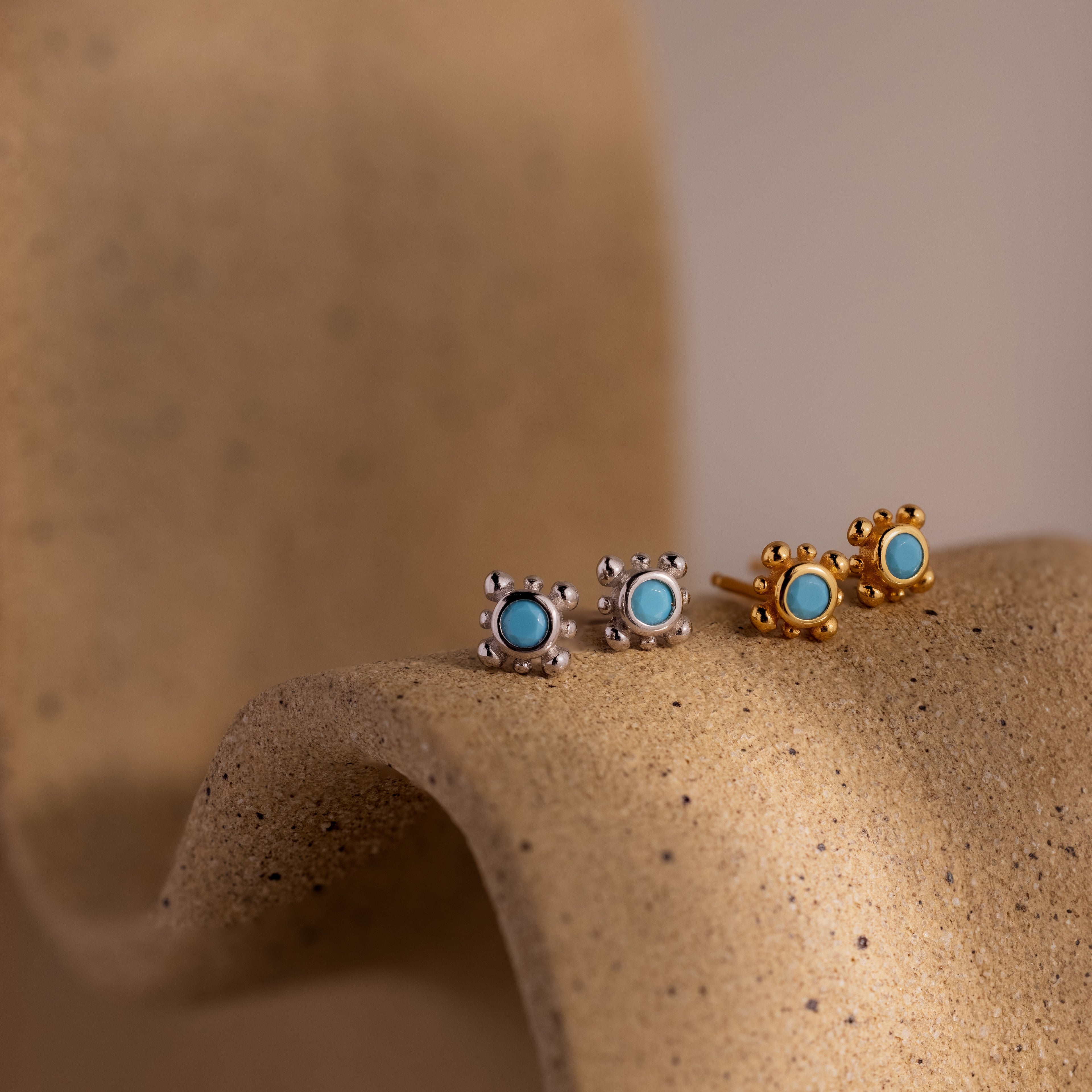 Two pairs of turquoise gemstone stud earrings in gold and silver on a textured beige surface