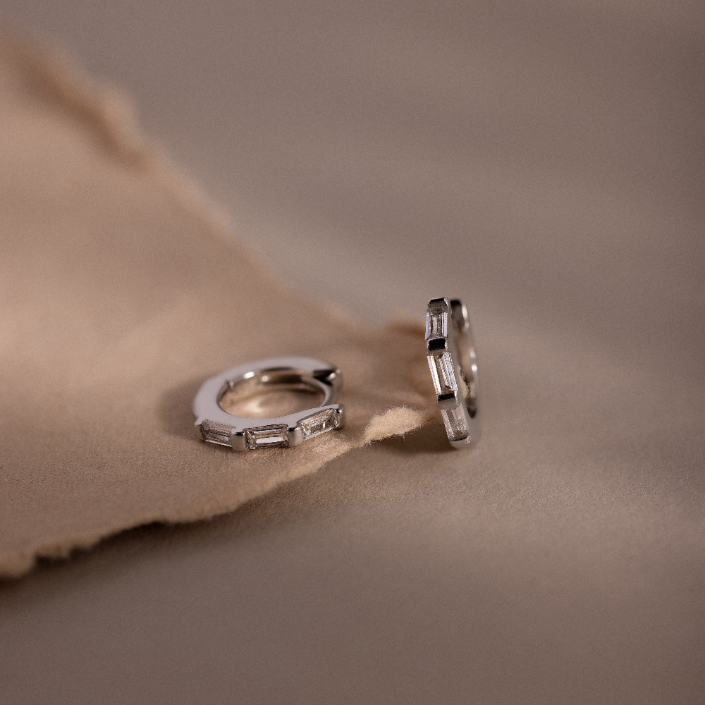 Pair of silver diamond baguette hoop earrings on a soft brown fabric background