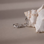 Silver diamond cluster hoop earrings on a sandy surface with a shell in the background