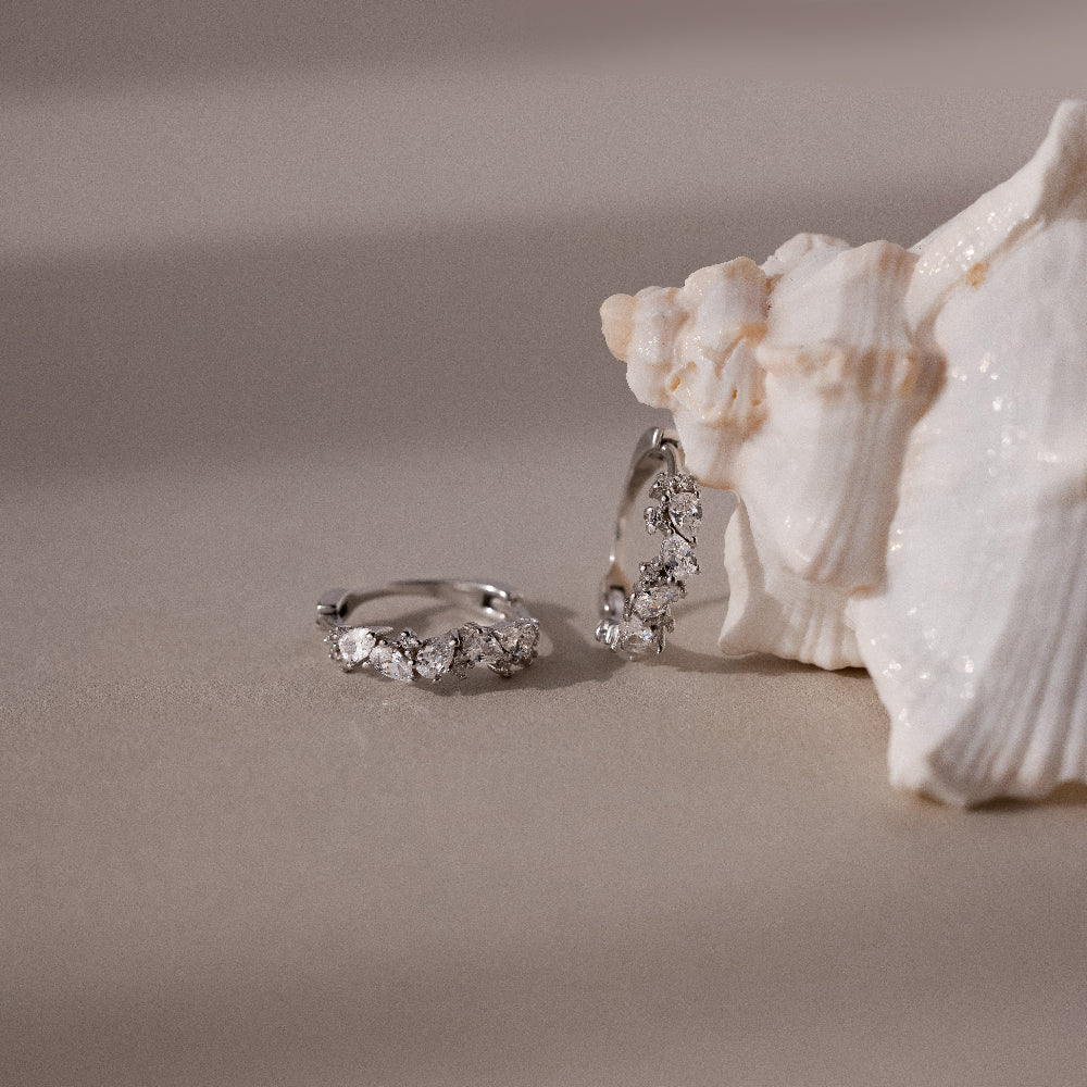 Silver diamond cluster hoop earrings on a sandy surface with a shell in the background