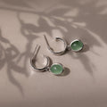 Silver stud hoop earrings with green jade gemstones on a beige surface with leaf shadows