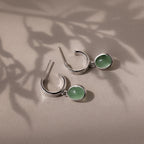 Silver stud hoop earrings with green jade gemstones on a beige surface with leaf shadows
