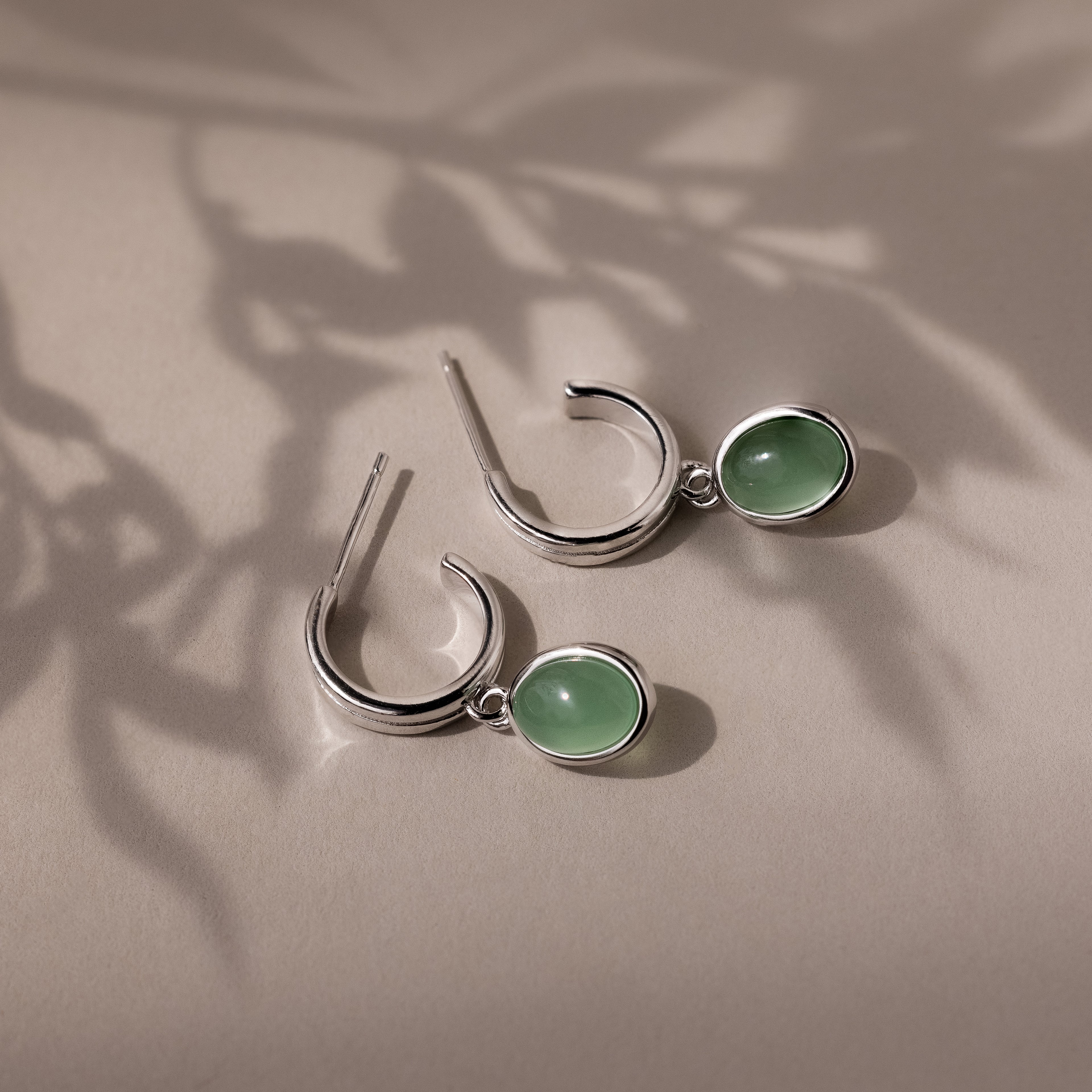 Silver stud hoop earrings with green jade gemstones on a beige surface with leaf shadows