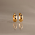 Bold Hammered Huggies in 18K Gold with a textured finish and abstract black designs are displayed on a reflective, neutral-colored surface.